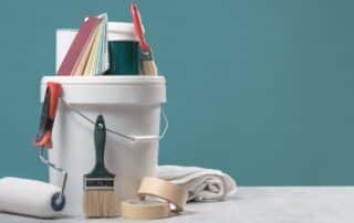 A bucket and various painting supplies, including brushes and tape, sit in front of a blue wall.