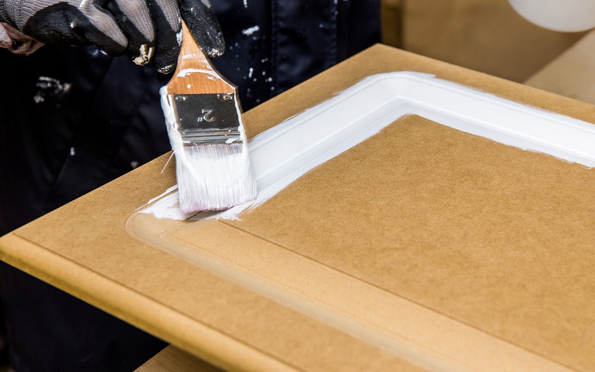 Repainting kitchen cabinet doors with white chalk paint indoors at home. Giving old kitchen new look concept. Hand holding a paint brush tool with paint against old cupboard door.