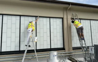 Two professional painters on ladders applying fresh exterior paint to a commercial building's upper facade.